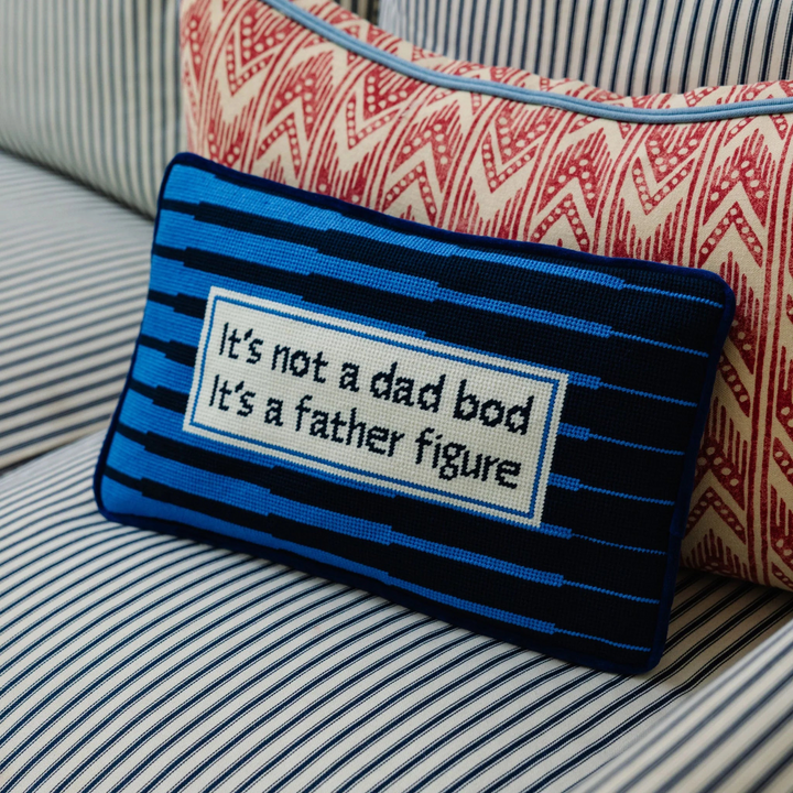 Ileana Makri Father Figure needlepoint pillow with blue striped design and witty text on a patterned sofa