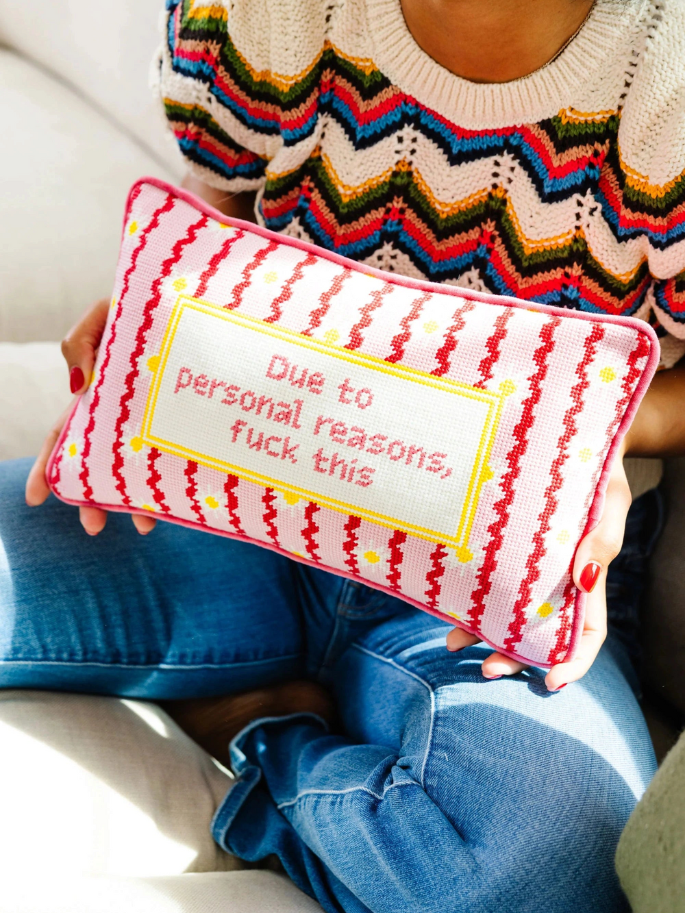 Ileana Makri needlepoint pillow with pink striped pattern and text held by a woman in a colorful knit sweater.