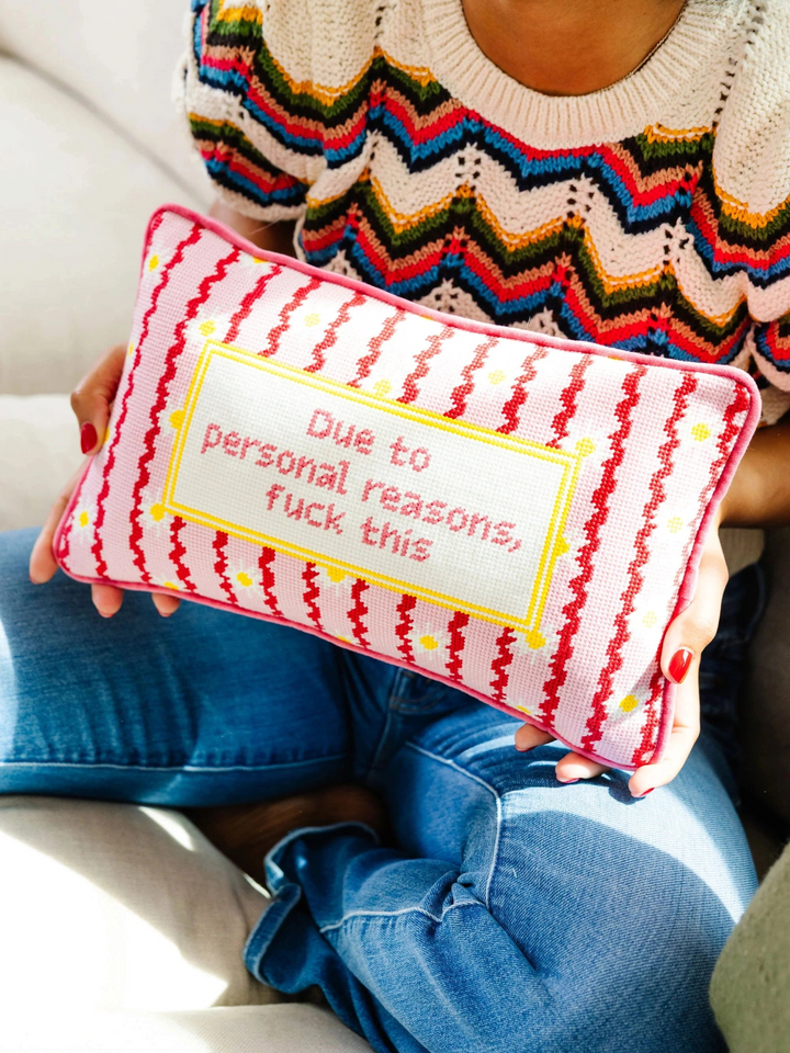Ileana Makri needlepoint pillow with pink striped pattern and text held by a woman in a colorful knit sweater.