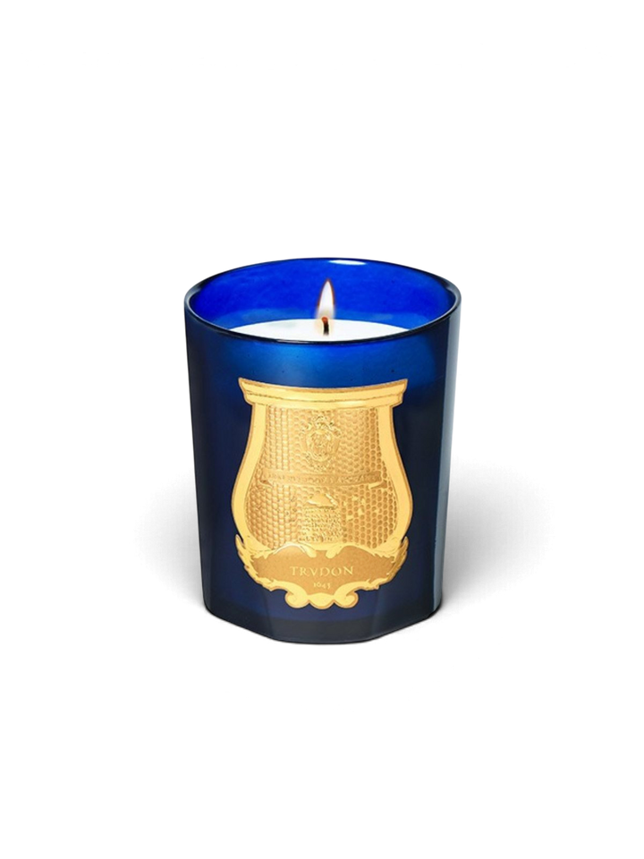 Ileana Makri Trudon Ourika candle in a deep blue glass vessel with a gold embossed emblem and a lit wick.