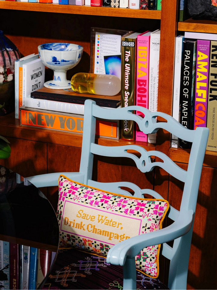 Ileana Makri Drink Champagne needlepoint pillow with colorful embroidery on a light blue wooden chair in a library
