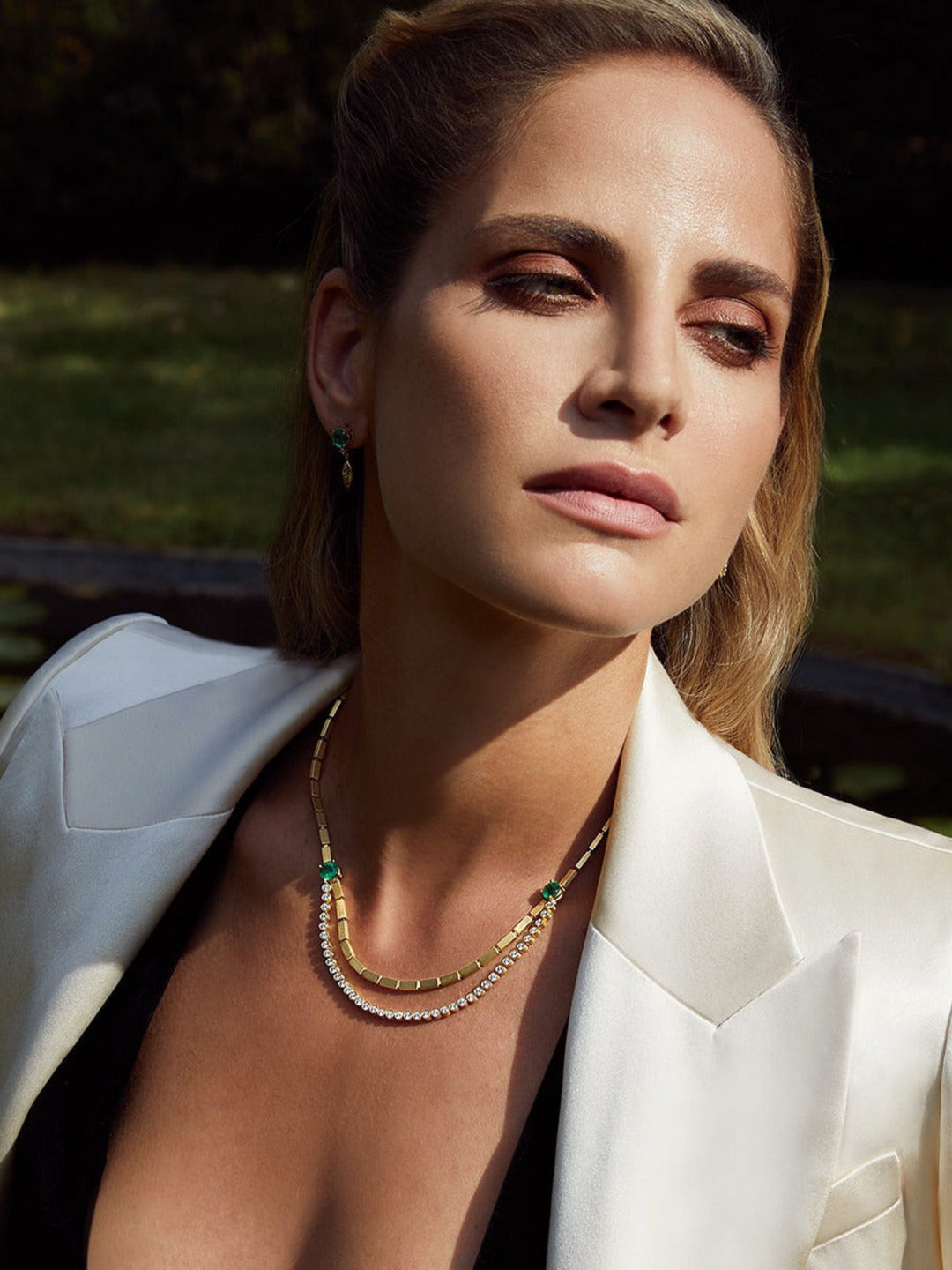 Ileana Makri Double Stream necklace in 18K yellow gold with emeralds and diamonds worn by a woman in a white blazer.
