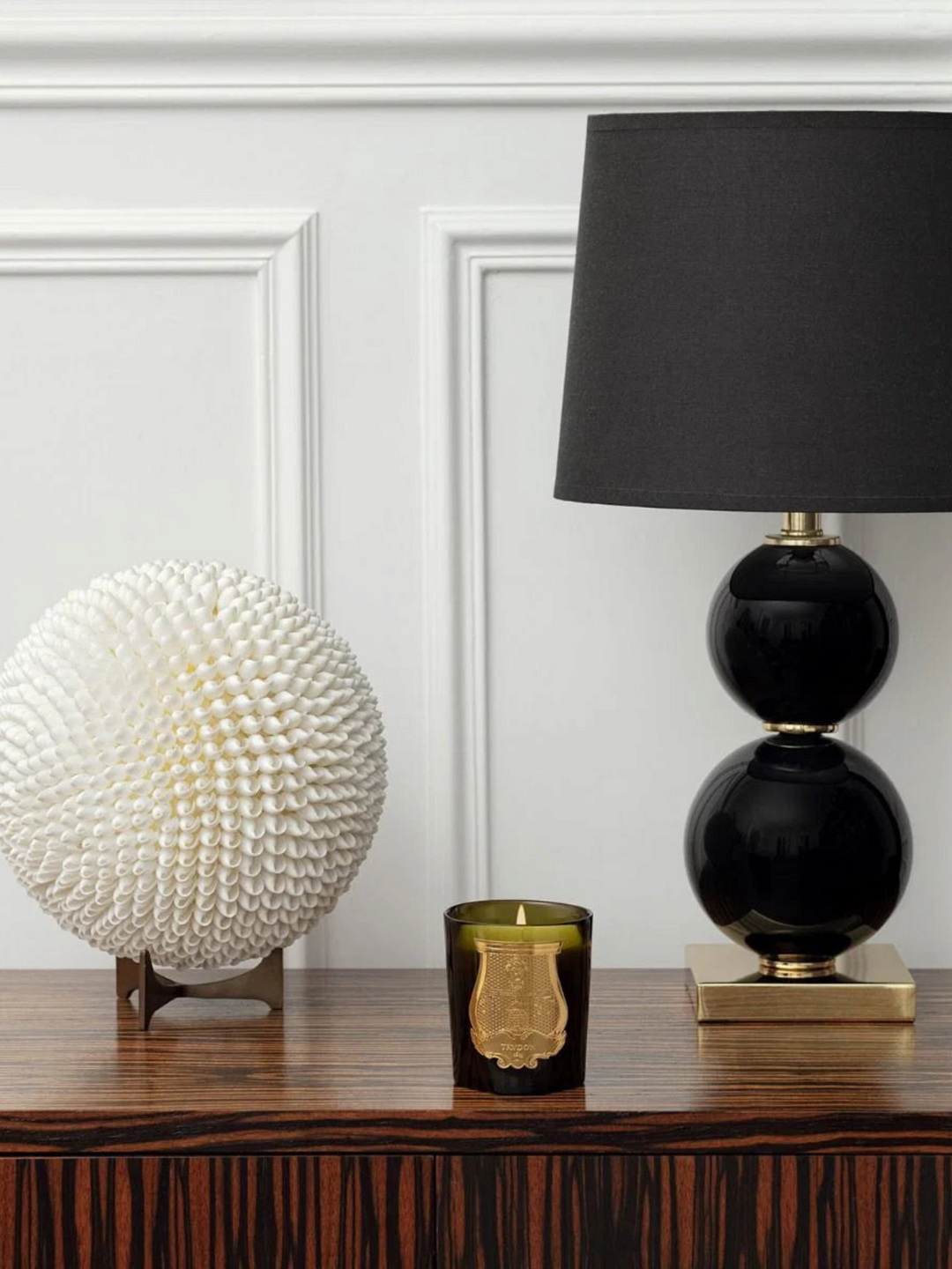 Ileana Makri Dada Tea & Vetiver scented candle in a dark green glass jar placed on a wooden table next to a lamp.
