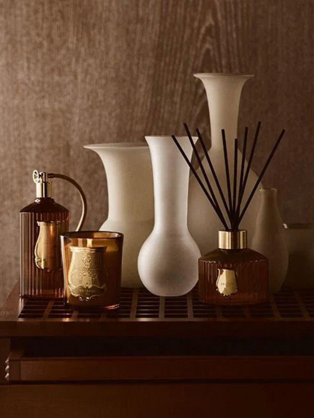 Ileana Makri Cire Trudon Altair candle and reed diffuser set with amber glass and gold accents on a wooden shelf