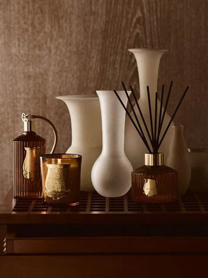 Ileana Makri Cire Trudon Altair candle and reed diffuser set with amber glass and gold accents on a wooden shelf