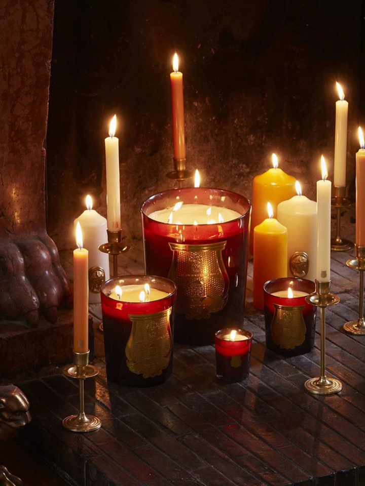 Ileana Makri Cire beeswax candles in amber glass vessels with gold emblems, arranged with lit taper candles on a hearth