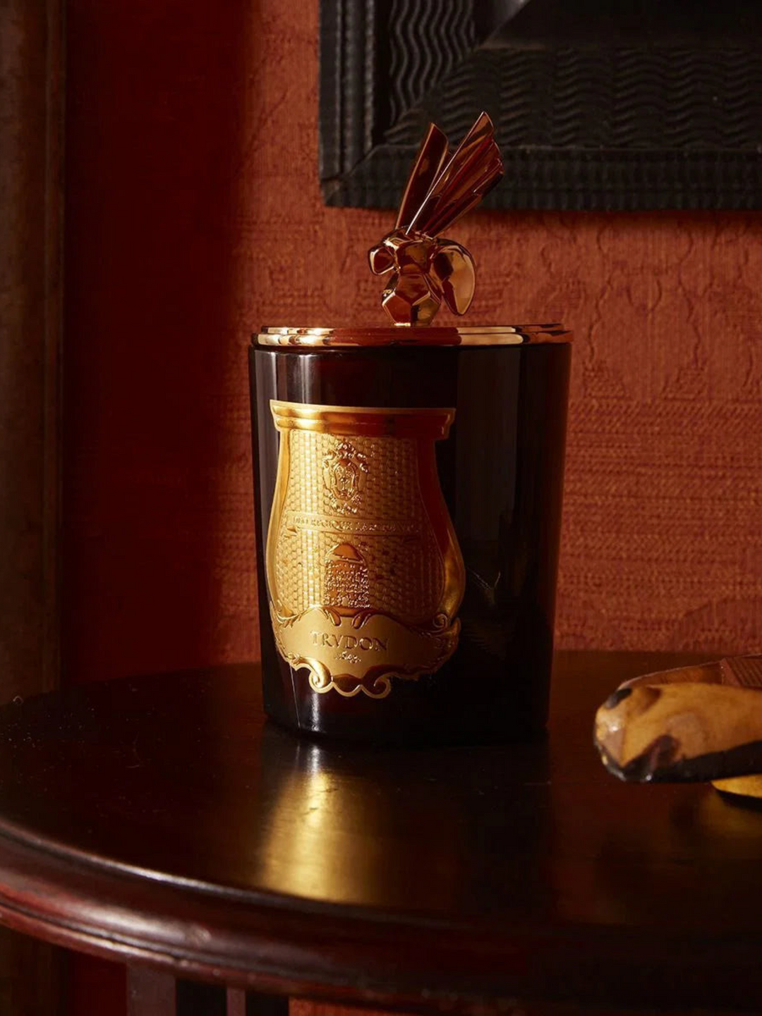 Ileana Makri Trudon Cire beeswax candle in amber glass with a gold bee lid and ornate gold emblem on a dark table.